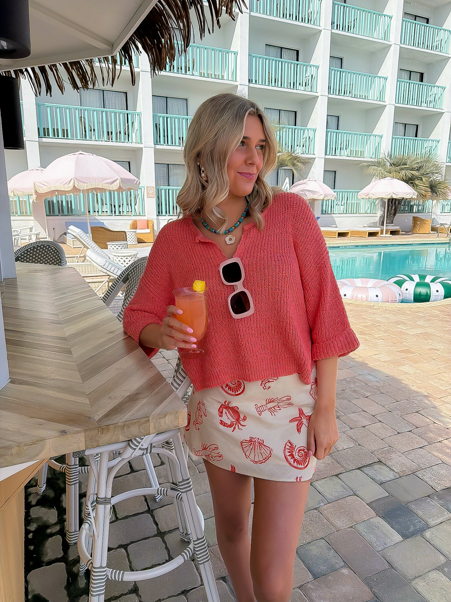Sally Red Seashell Printed Mini Skirt styled with a coral knit top at a poolside bar. Coastal-chic poppy red shell print perfect for summer getaways.
