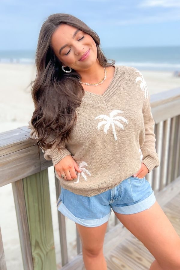 Palm Desert V-Neck Sweater in taupe with cream palm tree details, worn by a woman on a beach boardwalk. Relaxed fit, 100% cotton, perfect for vacation.