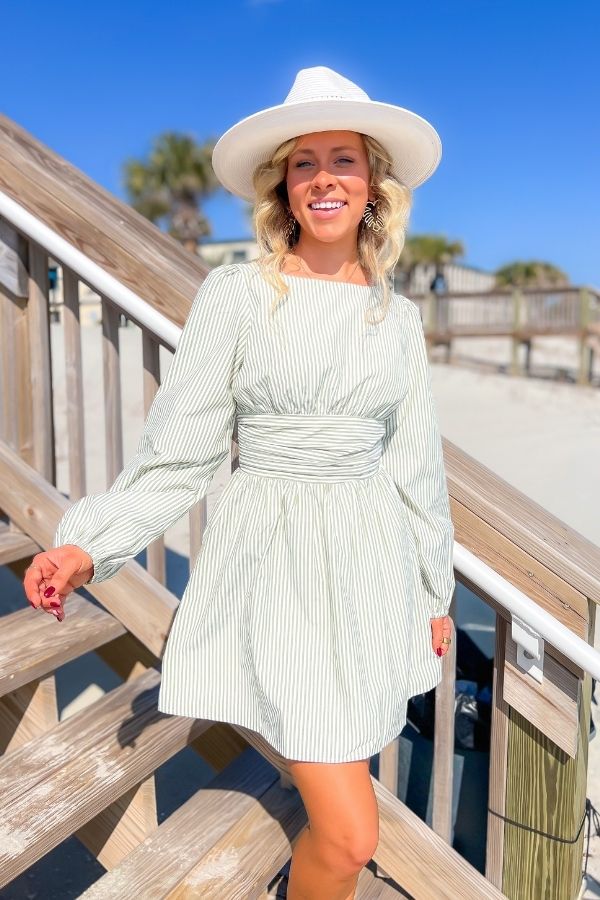 Elegant sage striped mini dress featuring an open back, tie waist, and long sleeves, styled with a white hat for a breezy resort-ready outfit.