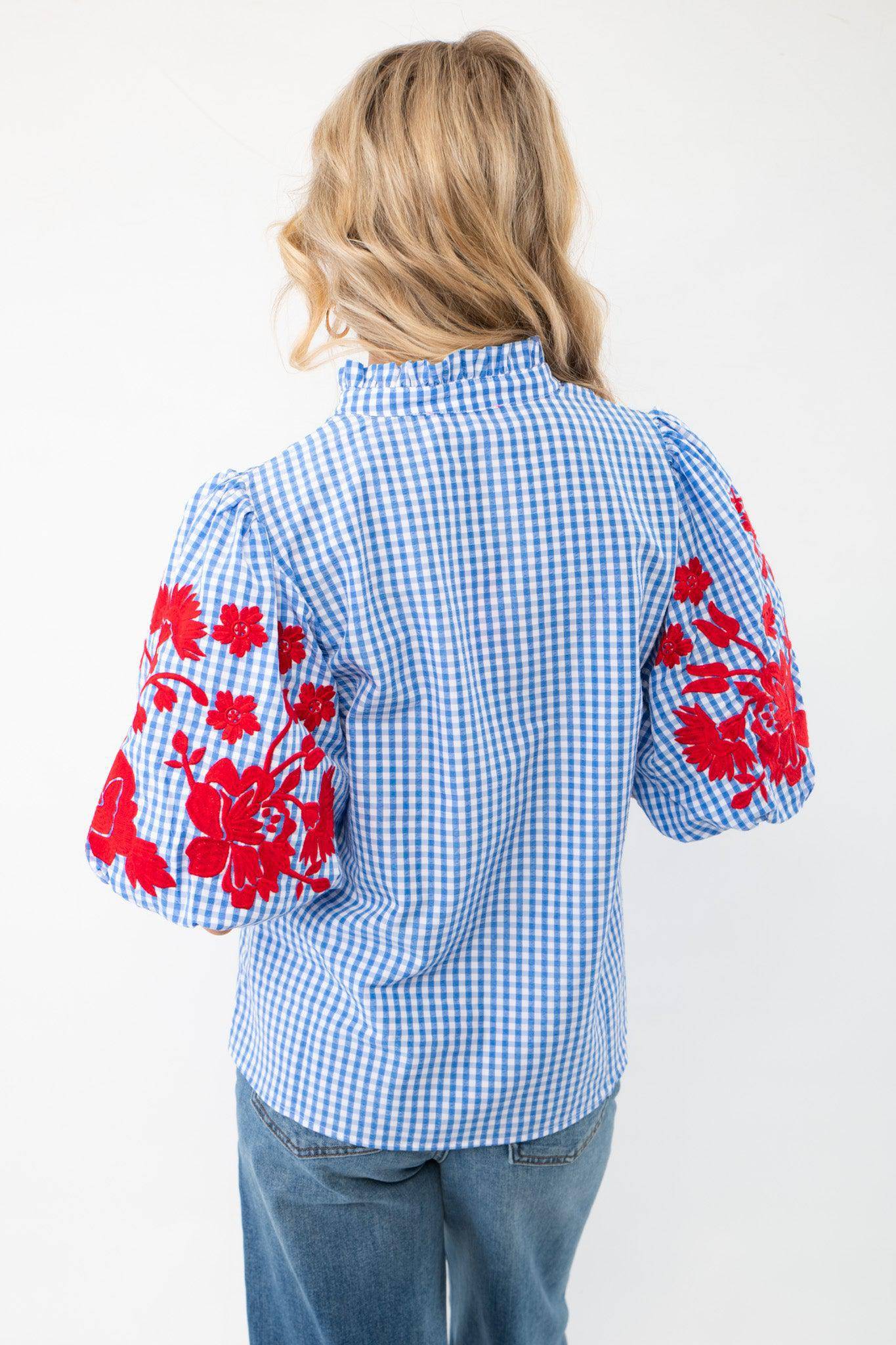 Back view of THML Blue Gingham Embroidered Top LM displaying puff sleeves with red floral embroidery and a ruffled neckline, styled with casual denim jeans.