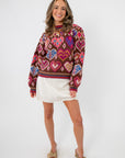 Chocolate Hearts Patterned Sweater – Cozy brown crewneck with colorful heart motifs, styled with a white skirt and heels for Valentine's Day.