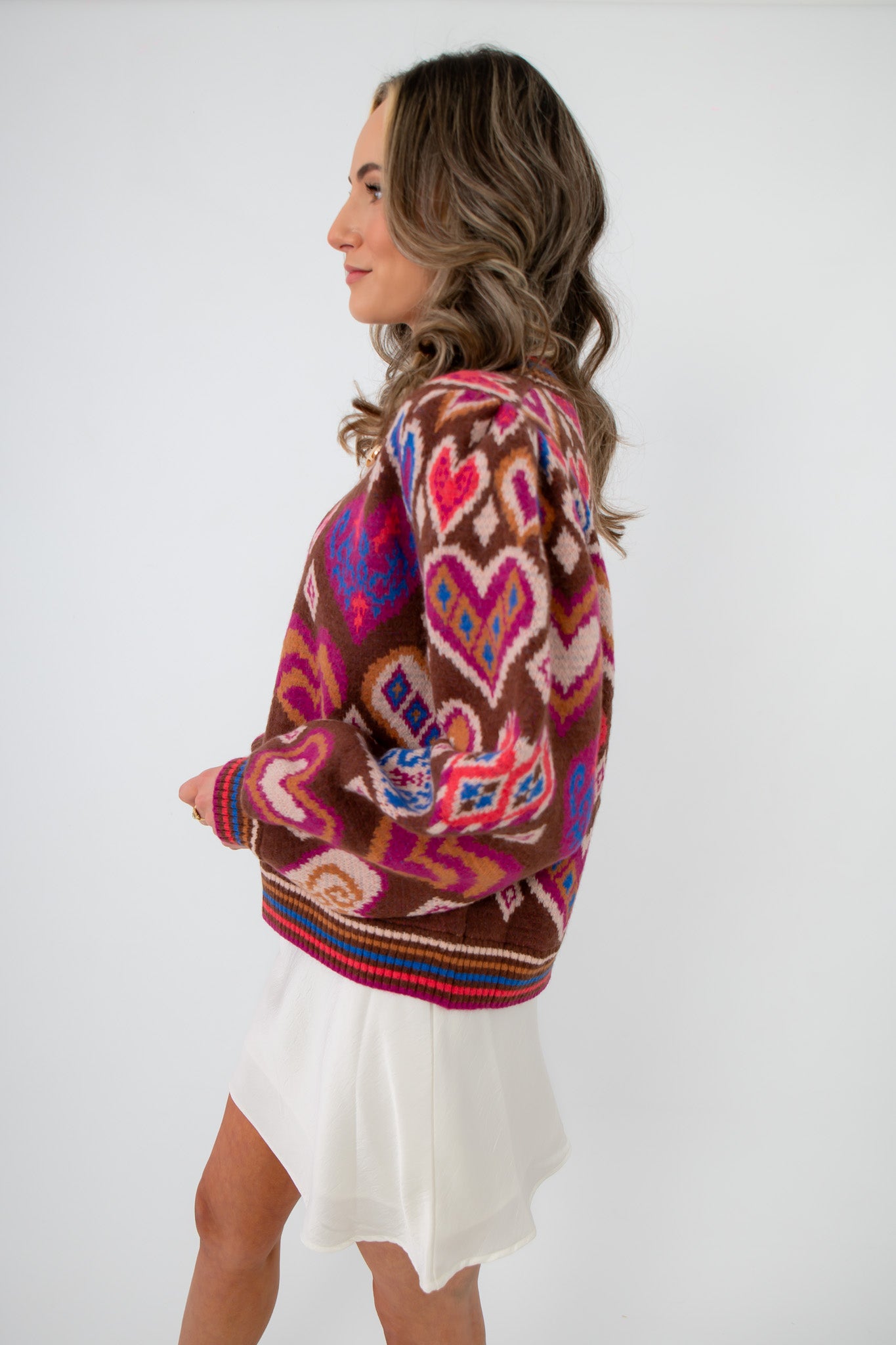 Side view of Chocolate Hearts Patterned Sweater featuring vibrant heart and geometric patterns on a brown base, paired with a white mini skirt.