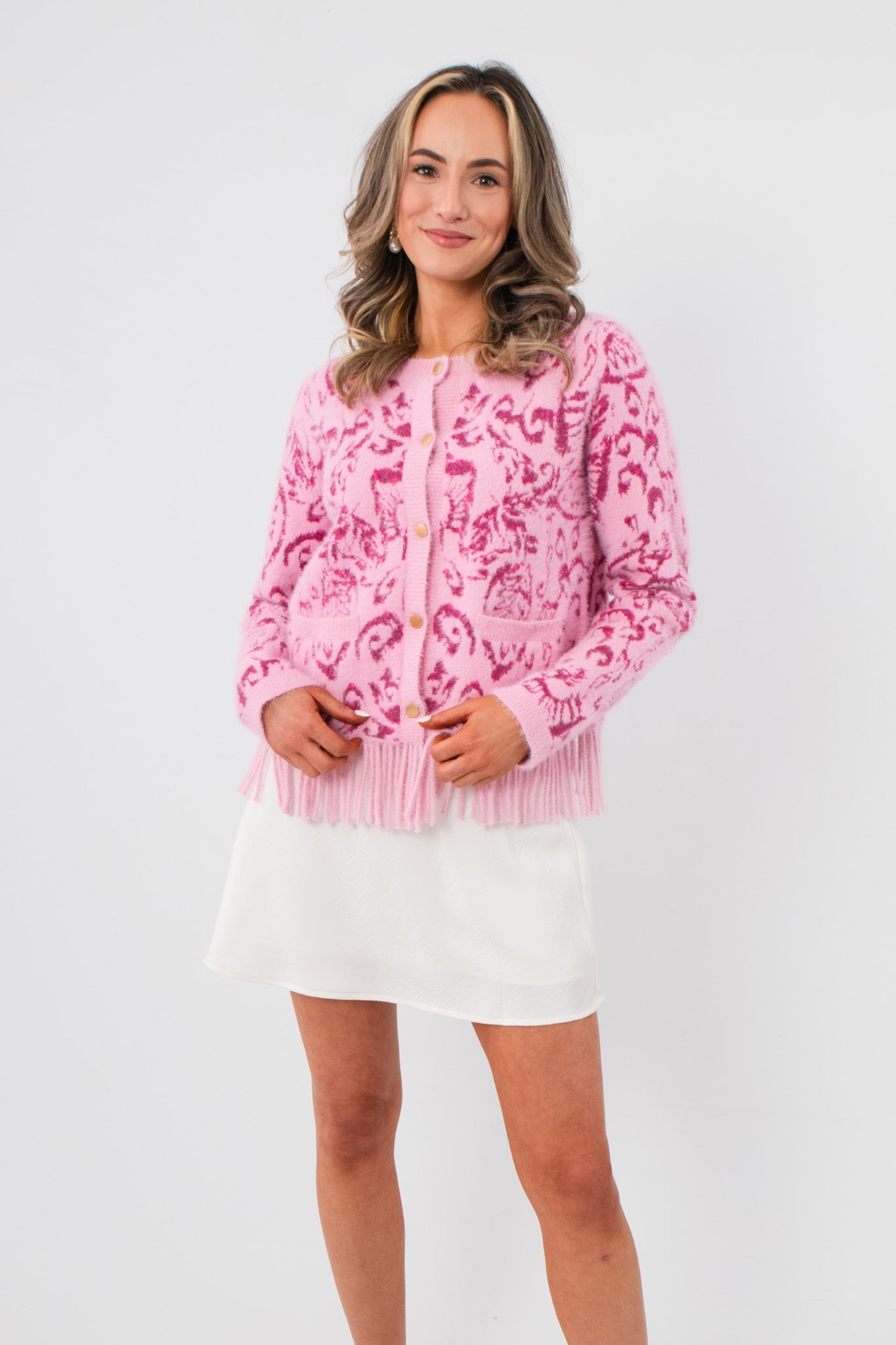 Something Sweeter Pink Jacquard Fringe Hem Cardigan with long sleeves, button front, and fringe detail, styled over a white mini skirt.