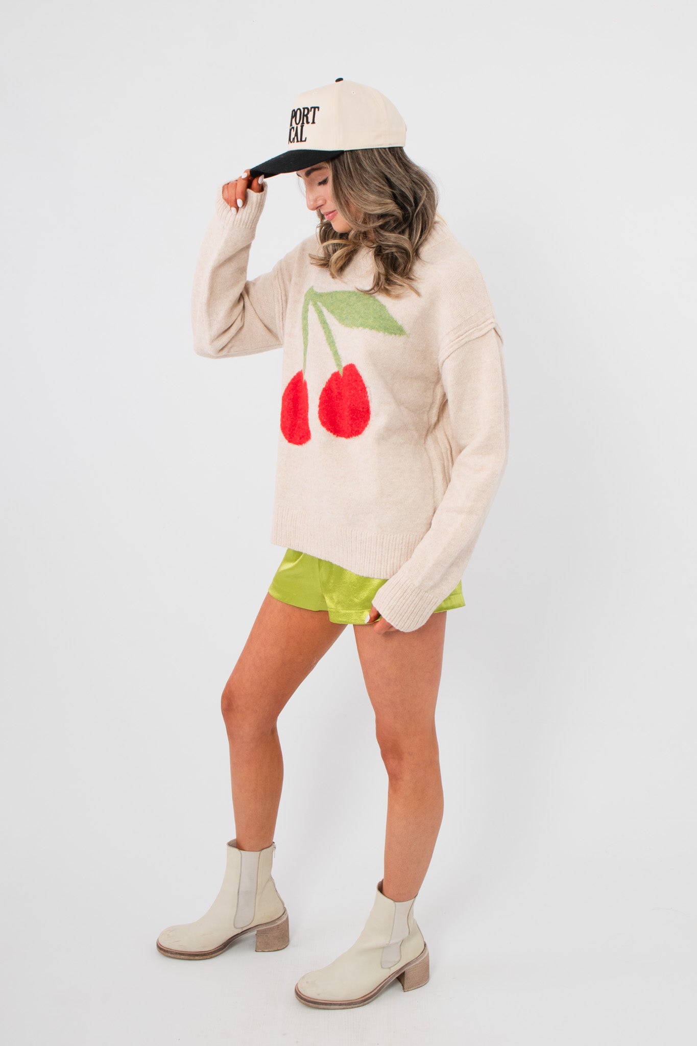 Side view of Cherry On Top Graphic Sweater – beige cozy knit with cherry print, styled for casual winter wear with boots and bright shorts.