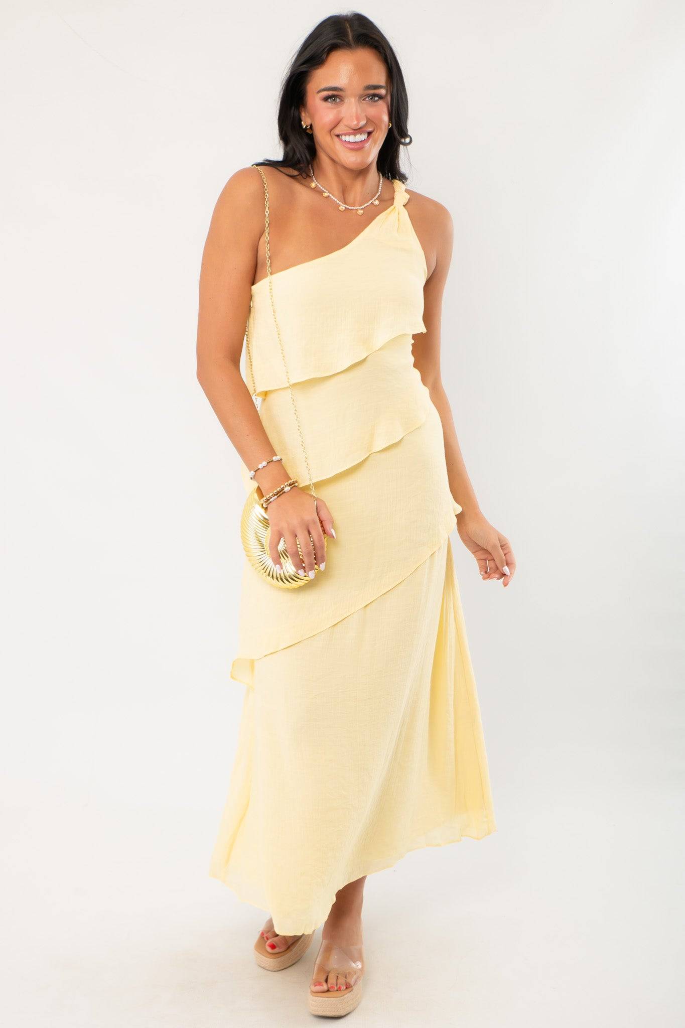 Andie Yellow Layered One Shoulder Maxi Dress styled with a gold clutch, ideal for weddings, parties, or vacation occasions.