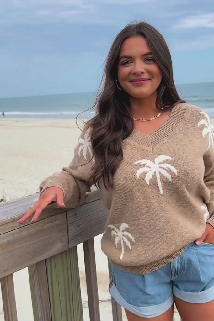 Taupe Palm Desert V-Neck Sweater with white palm tree design, styled casually with denim shorts on a beachside boardwalk. Lightweight and breathable.